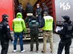Fotografia przedstawia ujęcie stojących tyłem funkcjonariuszy z napisem na kamizelce Służba Celna oraz funkcjonariuszy Straży Granicznej.  W tle wnętrze naczepy samochodowej w kolorze czerwonym, we wnętrzu naczepy stoją nielegalni  cudzoziemcy.