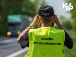 Funkcjonariusz w żółtej kamizelce z napisem Służba Celno-Skarbowa obserwuje przez lornetkę pojazdy.