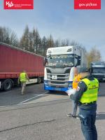 Na pierwszym planie funkcjonariusz w odblaskowej kamizelce stojący przy zaparkowanej ciężarówce i sprawdzający dokumenty, na drugim planie dwóch innych mężczyzn, z czego jeden w odblaskowej kamizelce.