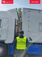 Funkcjonariusz w odblaskowej kamizelce stoi przed ciężarówką, drugi mężczyzna w odblaskowej kamizelce stoi na pojeździe między ciągnikiem a naczepą i przeprowadza kontrolę.
