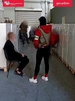 jeden z zatrzymanych mężczyzn siedzi na krześle, obok pilnujący go funkcjonariusz Służby Celno-Skarbowej, w tle funkcjonariusze Służby Celno-Skarbowej przeszukujący halę magazynową