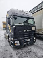 Widok na przód ciemnego ciągnika siodłowego, który stoi przy budynku przemysłowym.